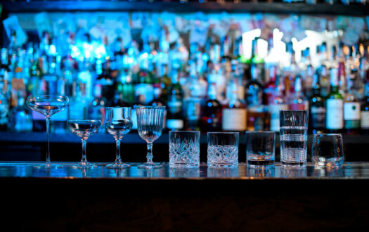 Several glasses of different forms stand on bar counter. Blue light on shelves with bottles of alcohol in background.